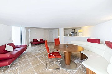 Bright, modern open-plan living space with red sofas, a dining area featuring a round wooden table, red chairs, and a white curved booth.