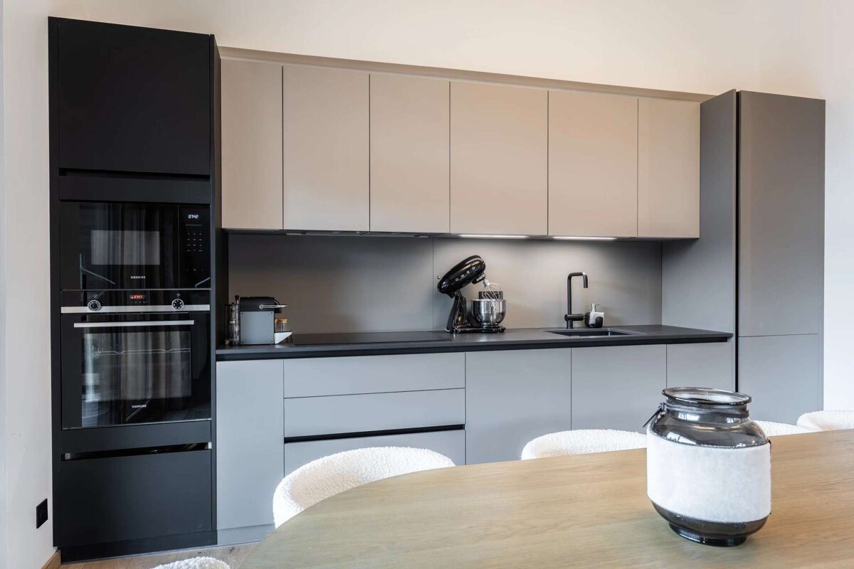 Sleek, modern kitchen with contrasting black, grey, and beige cabinetry, integrated appliances, and a minimalist countertop. A dining table is visible.