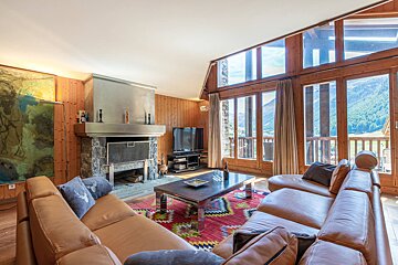 Cozy, wood-paneled living room with a stone fireplace, leather sofas, a colorful rug, and large windows offering a stunning mountain view.