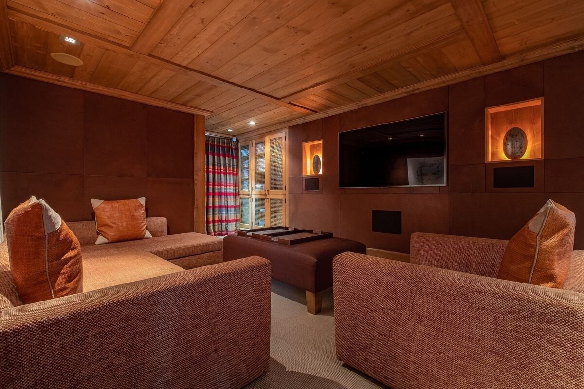 A cozy, chalet-style media room with wooden ceilings, brown walls, comfy orange sofas, a large TV, and decorative lighted niches.
