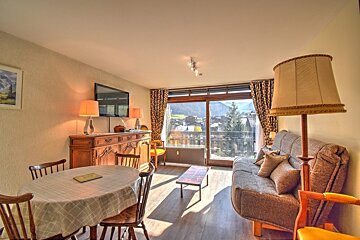 Sunny living/dining room with traditional decor, sofa, TV, dining table, and a balcony overlooking a charming village amidst mountains.
