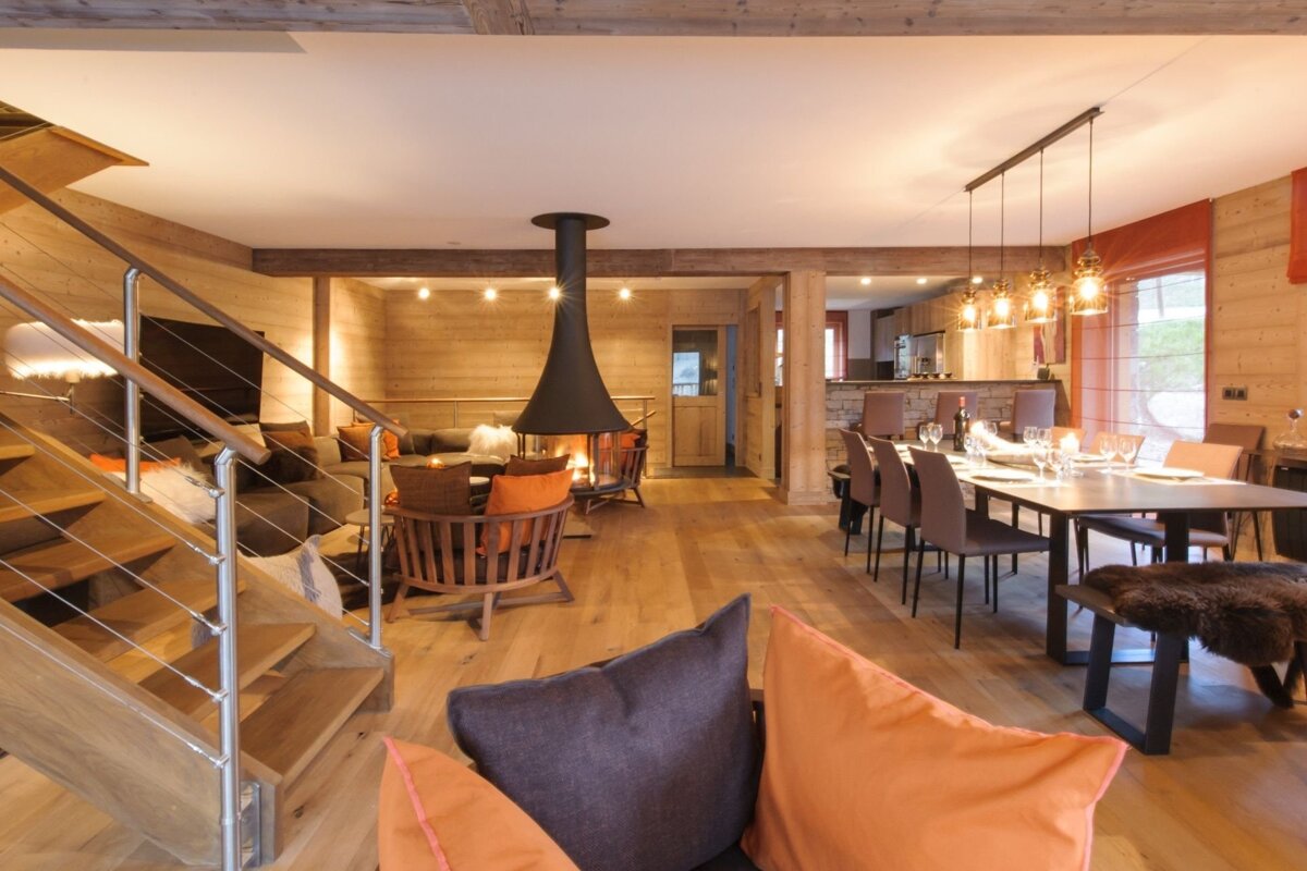 Cozy, open-plan chalet living room and dining area with a central modern fireplace, warm wood decor, and a sleek staircase.