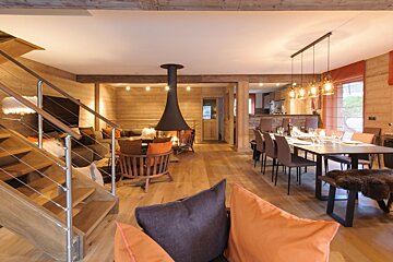Cozy, open-plan chalet living room and dining area with a central modern fireplace, warm wood decor, and a sleek staircase.