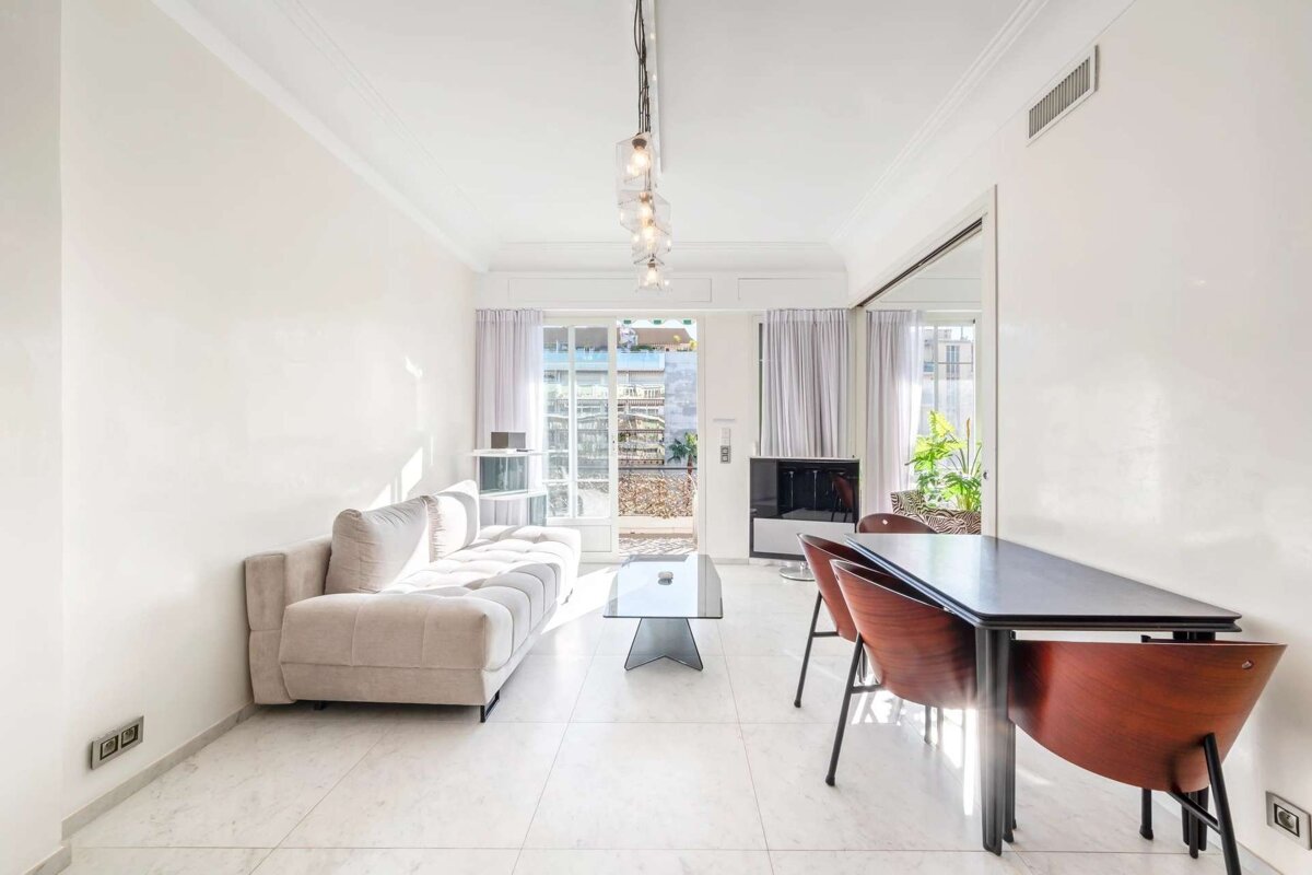 Bright, modern living/dining room with a light-colored sofa, glass coffee table, and a dining set. Glass doors open to a sunny balcony.