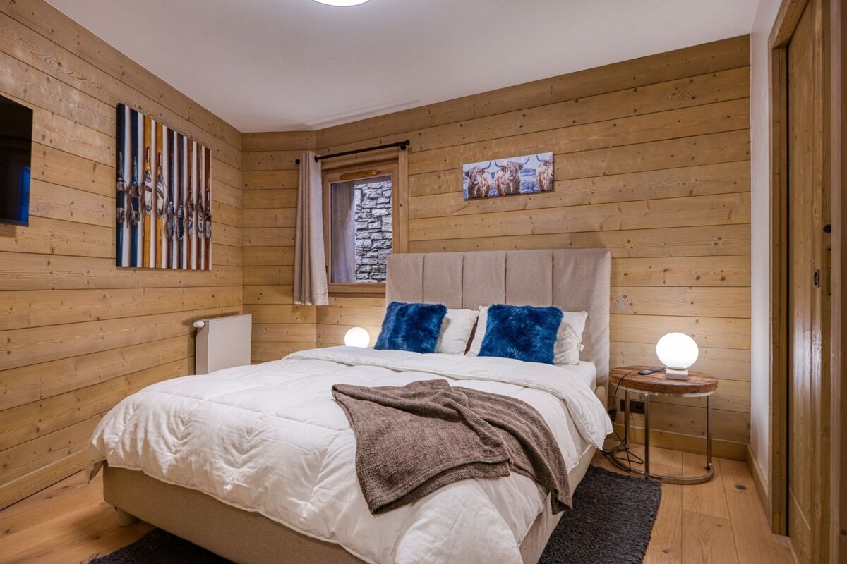 Cozy wood-paneled chalet bedroom with a double bed, bright blue accent pillows, and warm bedside globe lamps.