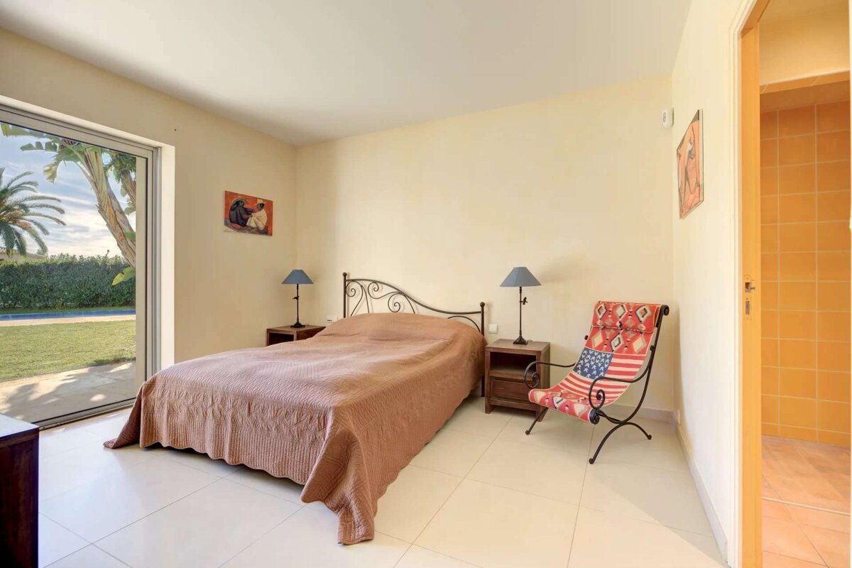 Bright bedroom with a bed, US flag chair, and large glass door opening to a garden with a pool and palm trees. Light walls and floors.