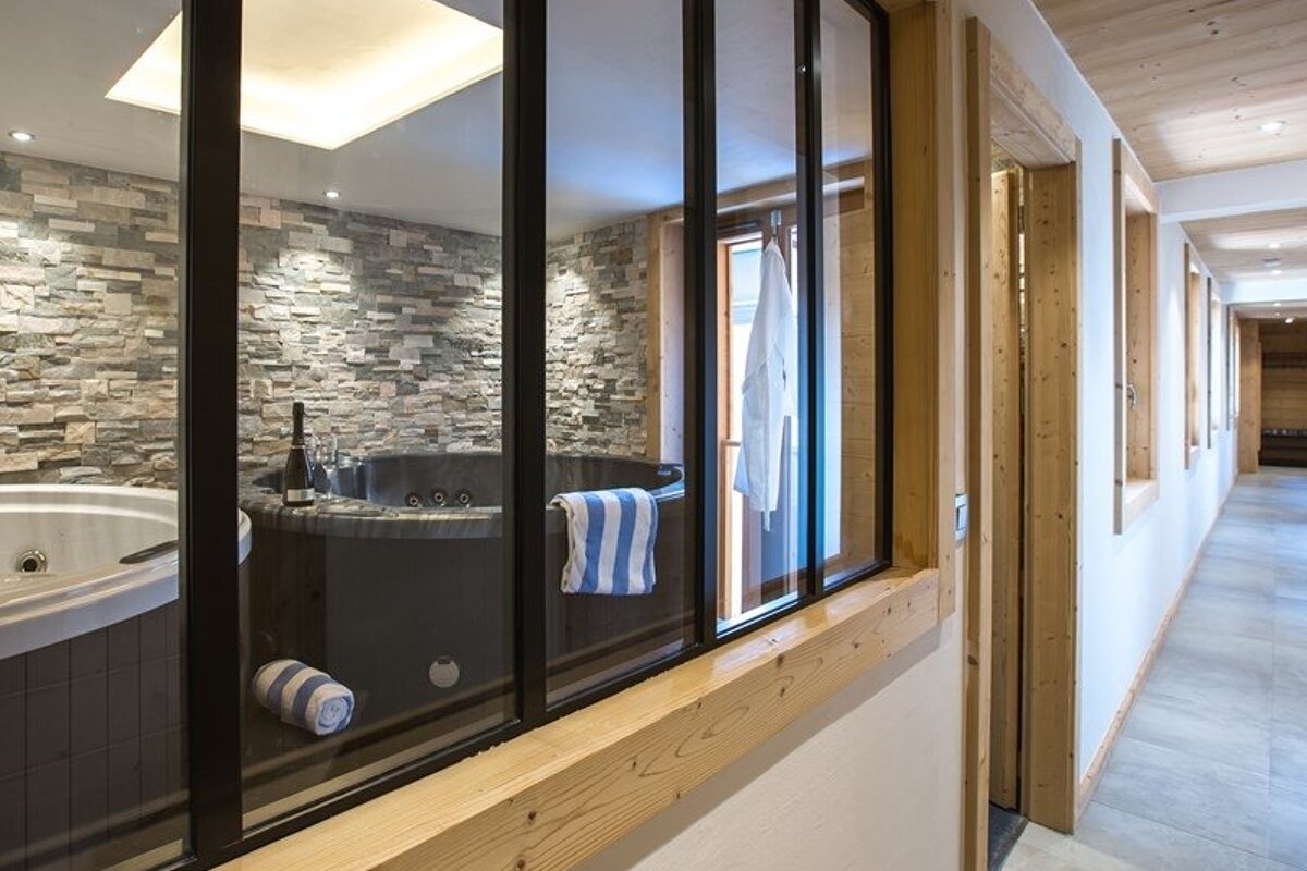 A view through a glass window of a modern indoor spa with a stone-walled hot tub, next to a bright hallway.