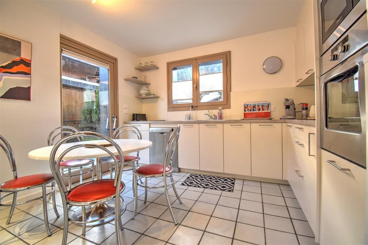 A bright kitchen with white cabinets, tiled floor, and a dining area with red chairs. Large window, integrated appliances.