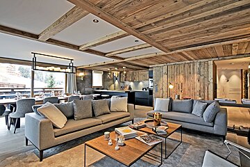 A luxurious open-plan chalet with rustic wood ceilings/walls, modern grey sofas, a dining area, and a kitchen, all with a snowy outdoor view.