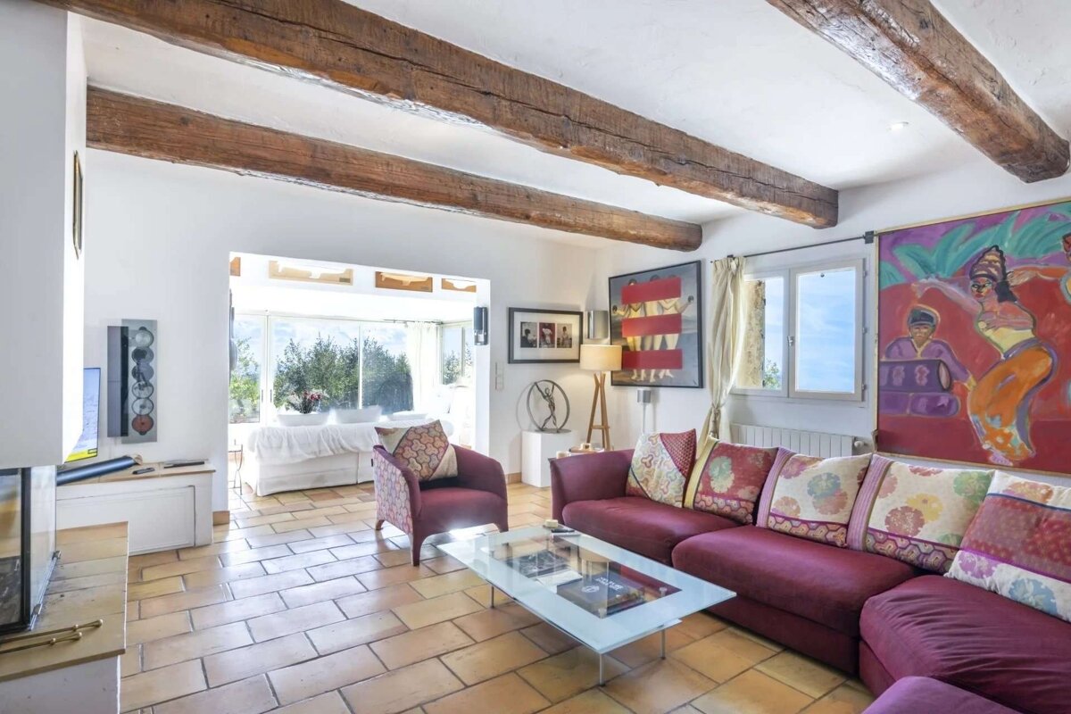 A rustic living room with exposed wooden beams, terracotta floor tiles, a maroon sofa, and colorful artwork. It's bright with a view outside.