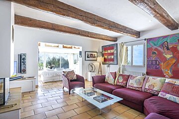 A rustic living room with exposed wooden beams, terracotta floor tiles, a maroon sofa, and colorful artwork. It's bright with a view outside.