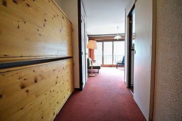 A red-carpeted hallway with light wooden paneling on the left leads into a bright room featuring a large balcony and a floor lamp.