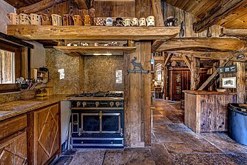 A cozy, rustic wooden kitchen with exposed beams, a vintage black stove, stone floor, and decorative mugs on shelves, exuding a cabin aesthetic.