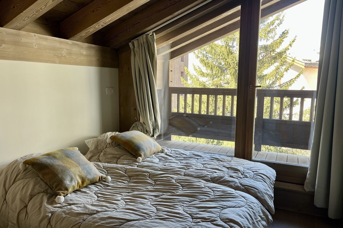 Cozy cabin bedroom with wooden ceilings, a quilted bed, and large glass doors looking out to a sunny balcony and pine trees.