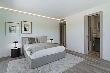 A modern, minimalist bedroom in white and grey. Features a comfy bed, bedside table, framed art, and an open door to a sleek bathroom.