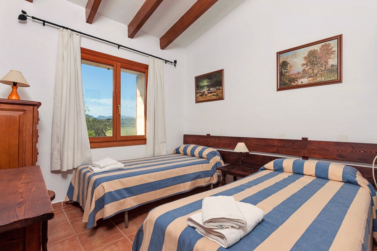 A rustic bedroom with two twin beds, blue/beige striped bedding, exposed wooden beams, and a scenic window view.