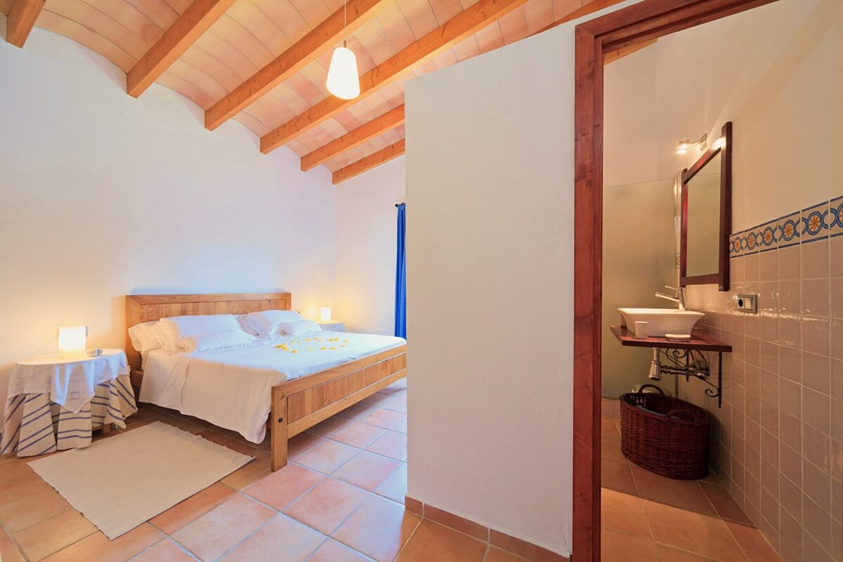 A rustic bedroom with a wooden bed, vaulted ceiling, and terracotta floor, featuring an adjacent bathroom with a sink visible through a doorway.