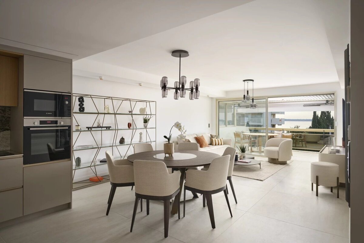 Modern open-concept dining and living area in neutral tones with a round table, geometric shelving, and access to a scenic balcony.