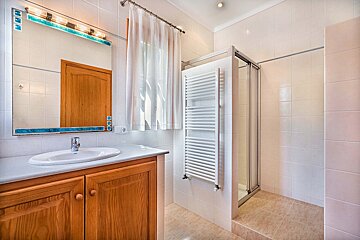 A tiled bathroom featuring a wooden vanity with an oval sink, a large mirror, a window with sheer curtain, a shower, and a white towel warmer.