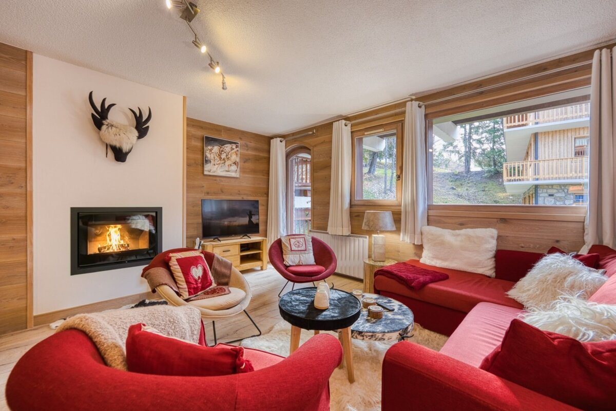 A cozy ski lodge living room with a lit fireplace, red seating, wood-paneled walls, and large windows looking out to other chalets.