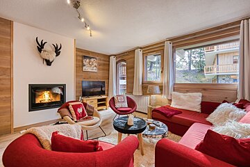 A cozy ski lodge living room with a lit fireplace, red seating, wood-paneled walls, and large windows looking out to other chalets.