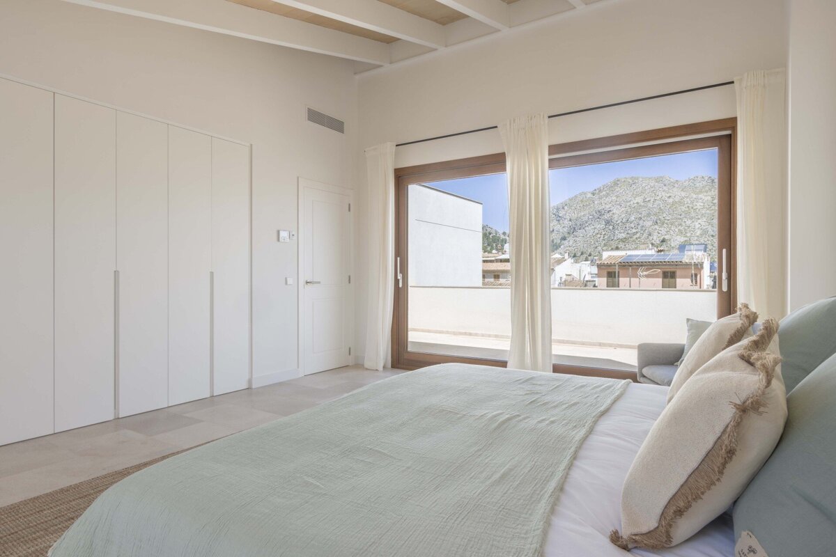 A bright, modern bedroom features a large bed with light bedding, white built-in wardrobes, and expansive windows offering a stunning mountain view.