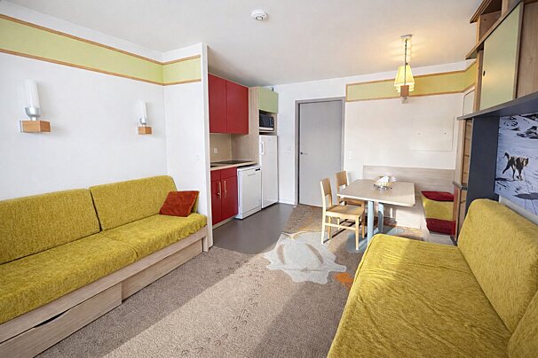 A small apartment interior featuring yellow sofas, a dining table, a small kitchen area with red cabinets, and light carpet.
