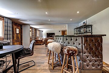 A cozy, rustic chalet-style interior featuring a tufted leather bar with fur stools, a dining area, and a comfortable living room beyond.