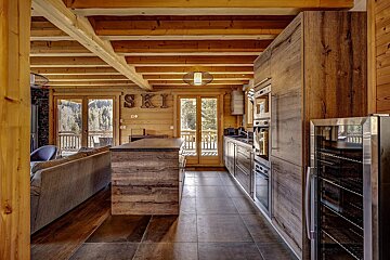 A cozy wooden cabin kitchen featuring exposed ceiling beams, a large island, modern appliances, and glass doors looking outside.