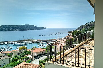 A sunny balcony overlooks a scenic Mediterranean harbor filled with boats and yachts, with lush green hills in the distance.
