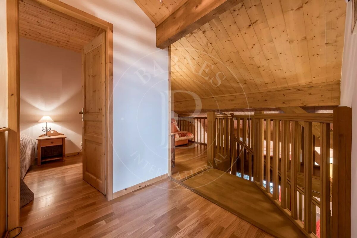 A cozy, rustic wooden interior of a home. Features a hallway leading to a bedroom with a lit lamp and a loft area with a wooden railing overlooking a lower floor.