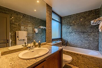 A modern bathroom with patterned granite walls, a sink, bathtub, and toilet. Features include a large mirror and a frosted window.