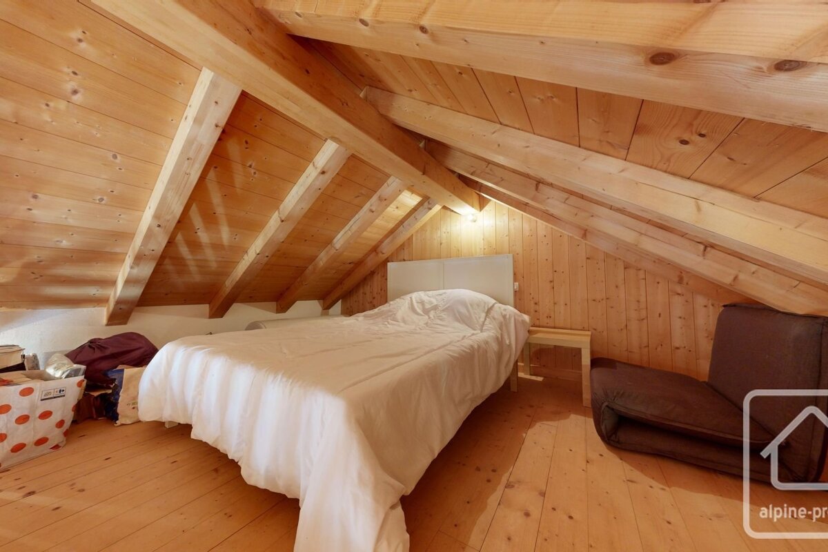 A cozy, rustic attic bedroom with warm wooden paneling on walls and ceiling, a white bed, and a brown lounge chair.