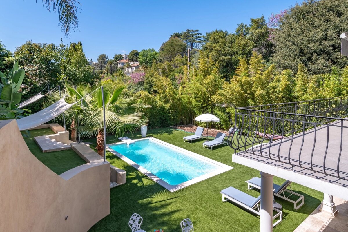 Elevated shot of a bright blue swimming pool on a green lawn with sun loungers, palm trees, and dense foliage, under a clear blue sky.