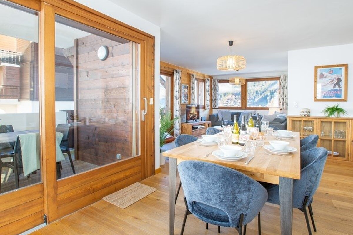 Bright alpine chalet dining room with a wooden table, blue chairs, and glass doors opening to a balcony.