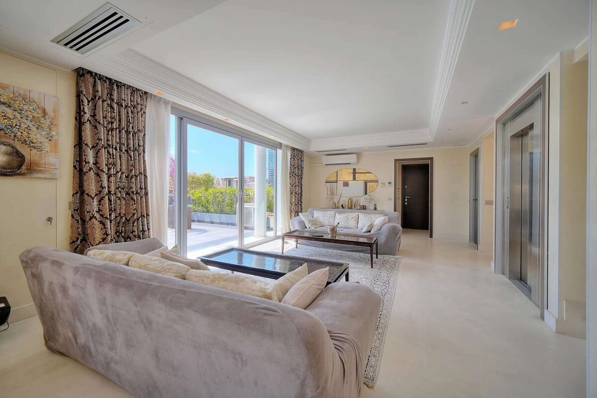 A spacious, modern living room in neutral tones with two sofas, large glass doors opening to a balcony, and a sleek, private elevator.
