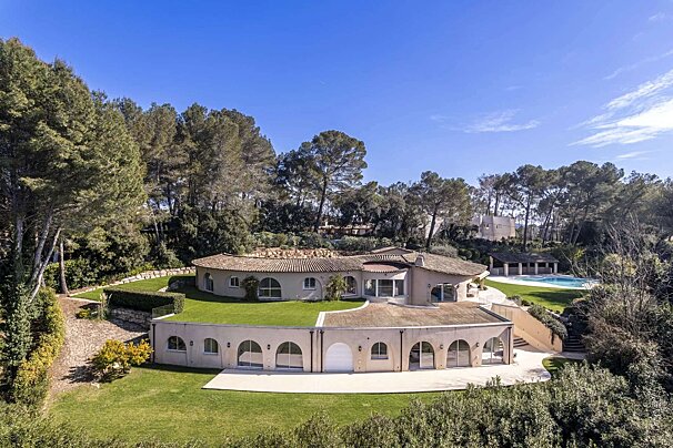 A large, curved luxury villa with terracotta roofs is nestled on a green, pine-covered hillside with terraced lawns and a distant pool under a clear blue sky.