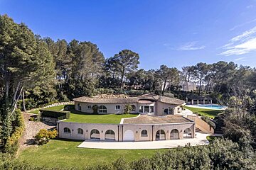 Aerial view of a luxurious, multi-level curved house nestled among lush green lawns and a dense forest under a clear blue sky, with a pool.