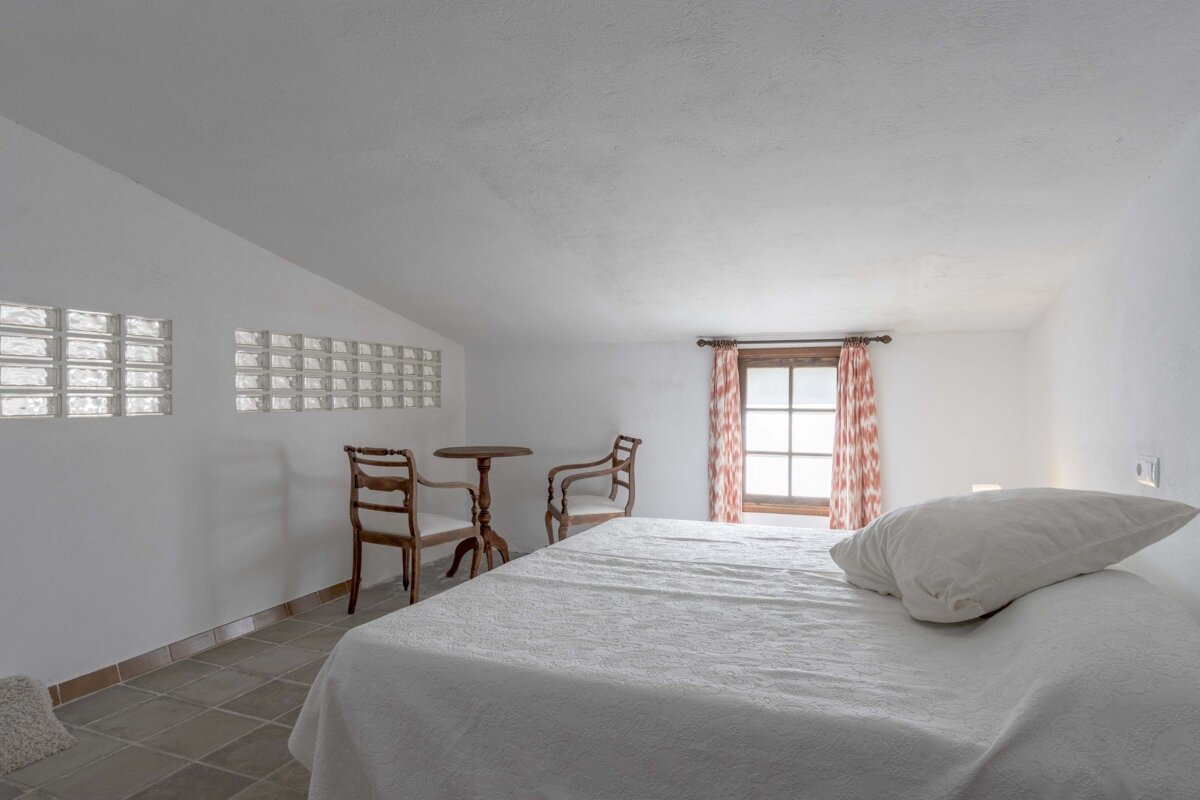 A simple, bright bedroom with white walls, a bed, a small table and two chairs, patterned window curtains, and decorative glass block windows.