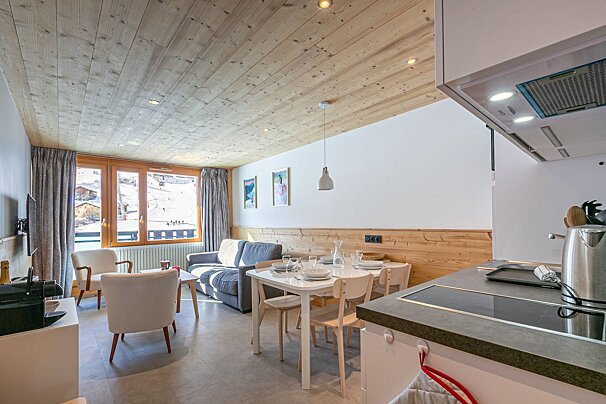 A cozy, modern open-plan living space with a dining area and kitchen, featuring wood-paneled ceiling/walls and a window overlooking ski slopes.