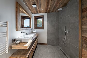 Modern rustic bathroom with a wooden ceiling, dual sinks, walk-in shower, and mountain views.