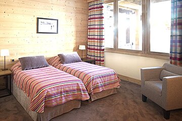 A cozy, wood-paneled twin bedroom with striped bedding, colorful curtains, a large bright window, and an armchair.