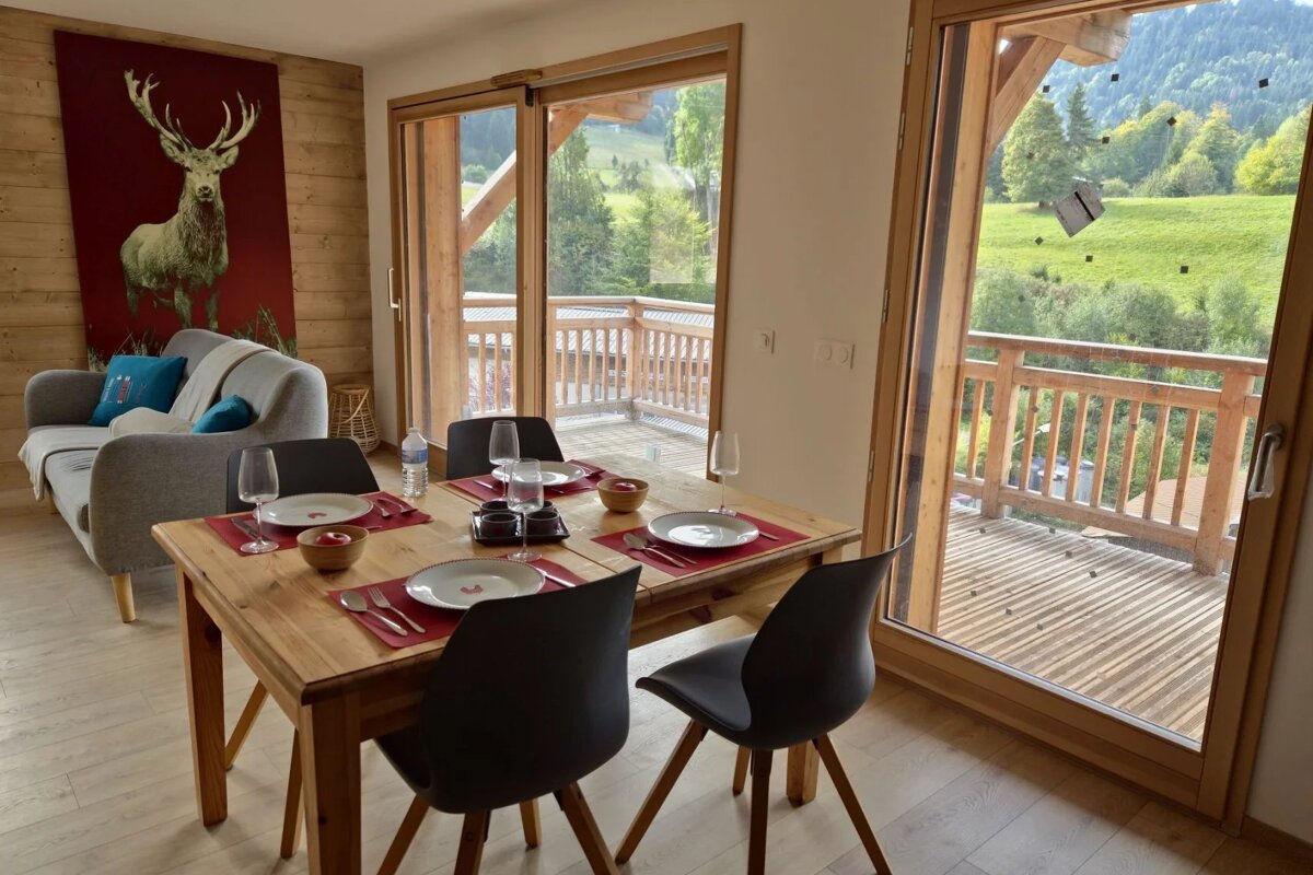 Cozy cabin dining area with a wooden table set for four, looking out through large glass doors onto a scenic green hillside.