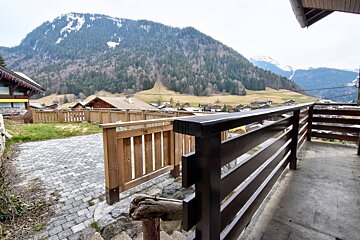 View of a mountain village and forested hills from a paved outdoor area and balcony with wooden railings in the foreground.