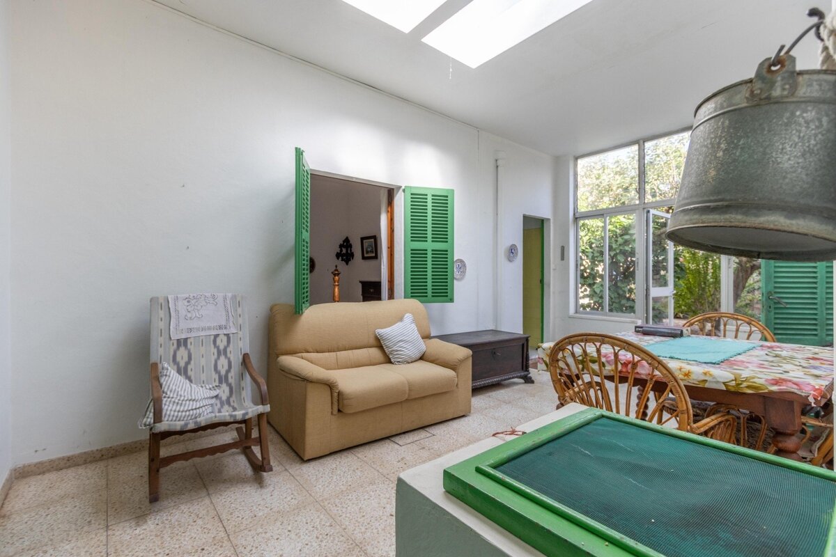 A bright, rustic sunroom with white walls, skylights, tan couch, rocking chair, and dining table. Green shutters, large windows, and a hanging metal bucket.