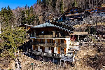 A multi-story chalet with wooden balconies on a forested mountain slope. Other chalets are visible on higher ground under a clear sky.
