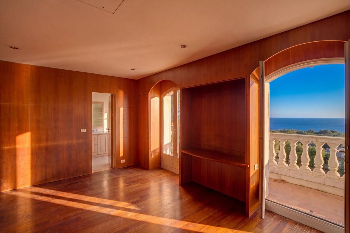 An empty, wood-paneled room with sunlight streaming in, featuring a built-in desk and an arched doorway opening to a balcony with a vast ocean view.