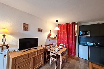A cozy, compact studio apartment featuring a dining area, TV, and a kitchenette. Wooden furniture and red curtains with a festive deer pattern complete the space.