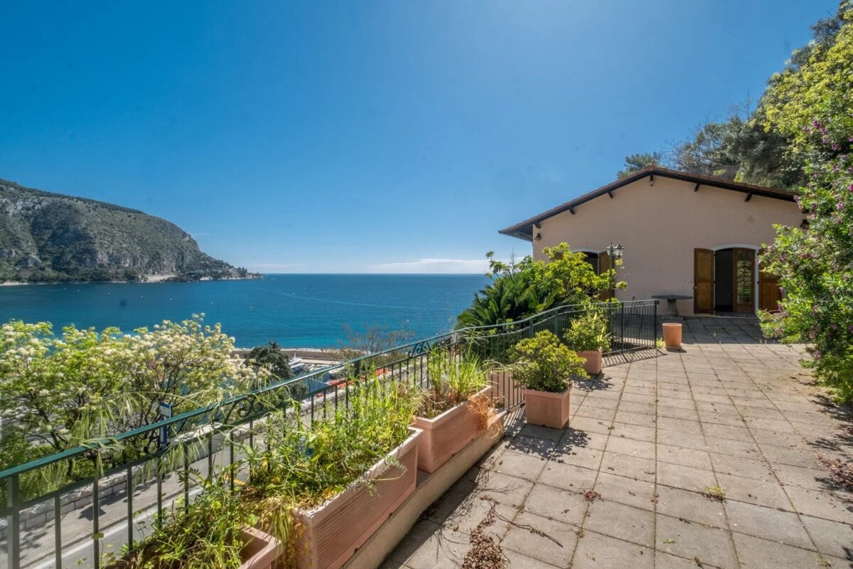 A sunny house patio with potted plants offers panoramic views of a deep blue bay, distant green mountains, and a clear sky.