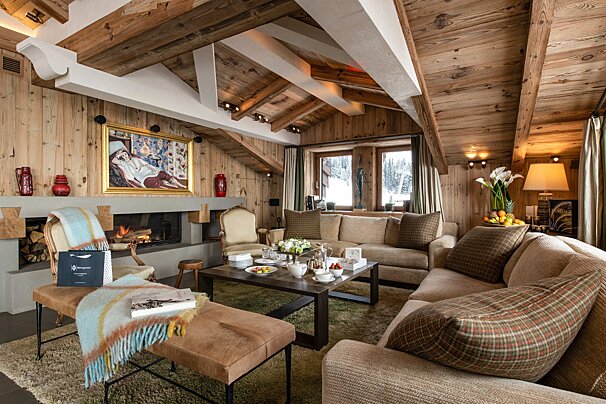 A warm, rustic chalet living room featuring wooden walls/ceiling, a fireplace, comfortable sofas, and a snowy mountain view.